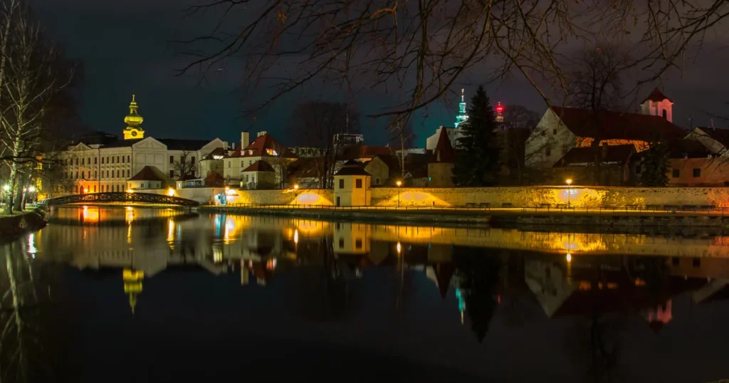 Noční pohled na historické centrum Českých Budějovic s osvětleným mostem a odrazy světel na hladině řeky.