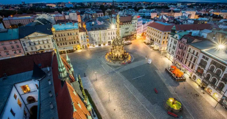 Vánoční trhy Olomouc 2025 – pohled na Horní náměstí s orlojem a Sloupem Nejsvětější Trojice ozdobeným vánočními světly.